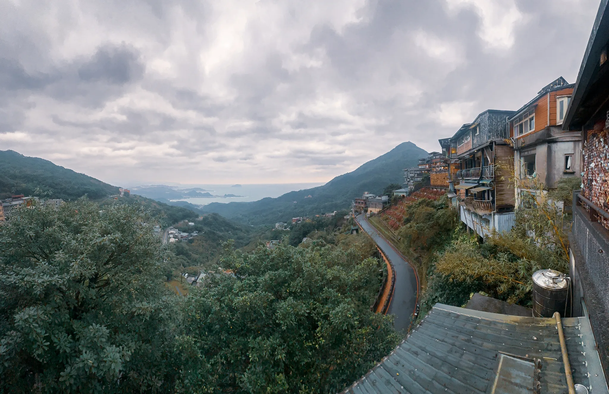 Teahouses stacked down a green hillside above a hairpin road, the sea and a line of headlands opening out under heavy cloud