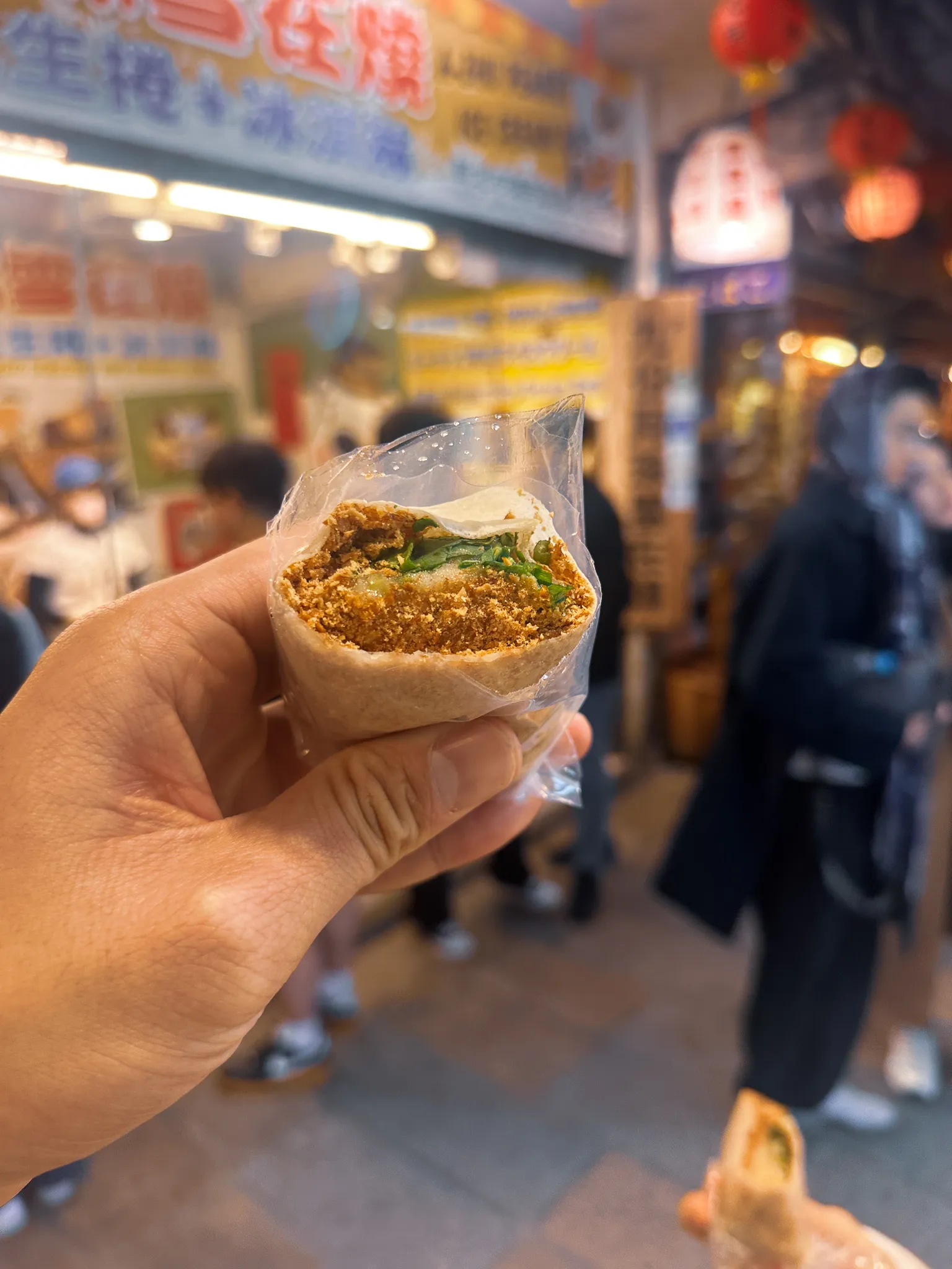 A peanut ice cream roll held up in its wrapper, shaved peanut brittle and coriander packed into the pancake, the stall's blue signs and red lanterns behind