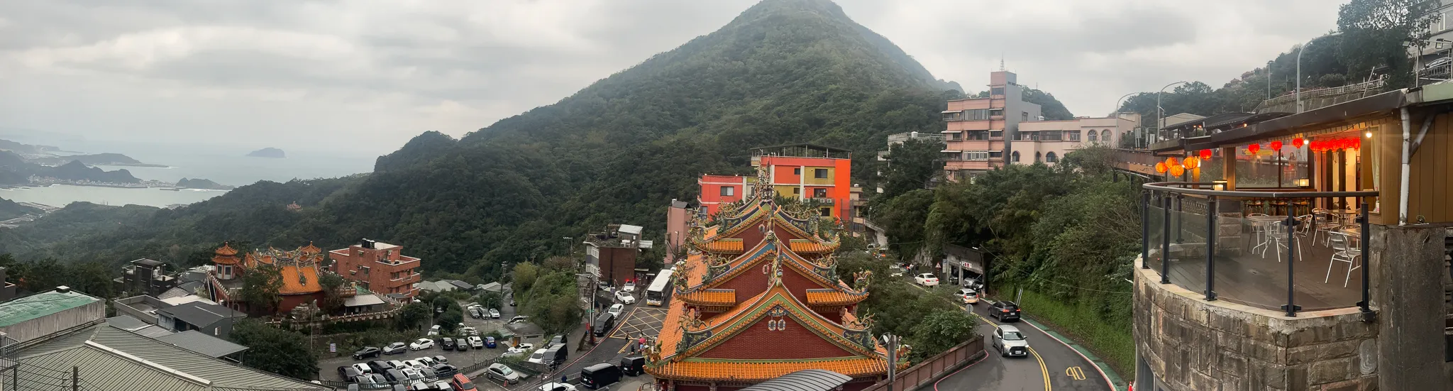 A wide look across the valley from a terrace, a temple's ornate tiled roof below, forested peaks behind it and the sea flattening out to the left