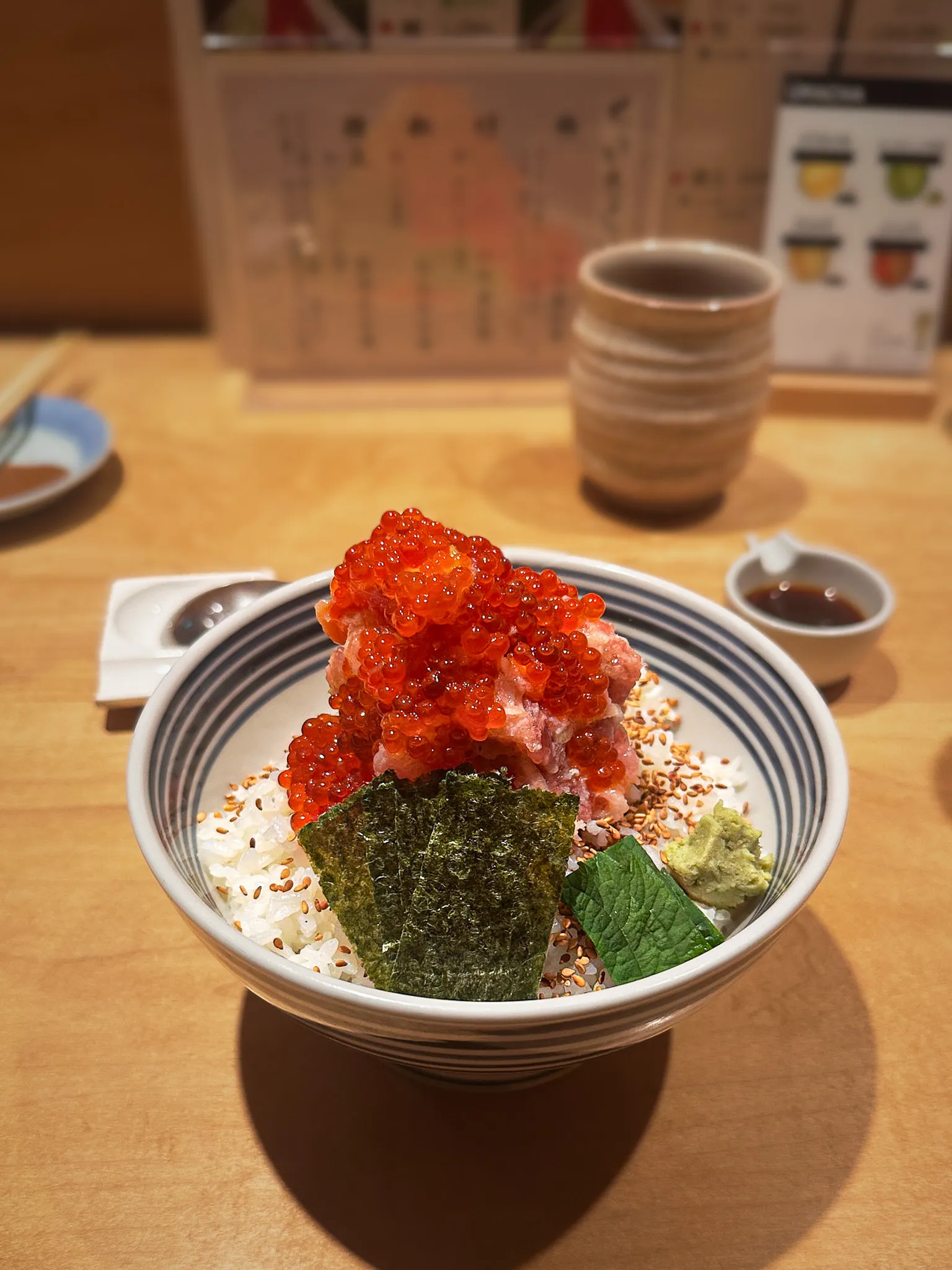 A rice bowl heaped with salmon roe and minced tuna, a sheet of nori and a dab of wasabi beside them, on a pale wooden counter