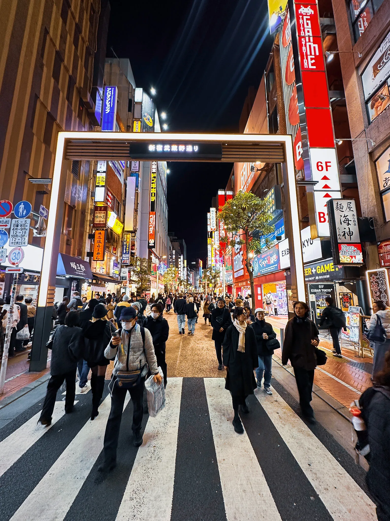 A lit gateway spanning a Shinjuku street at night, neon signs stacked up both sides and people crossing on the stripes beneath