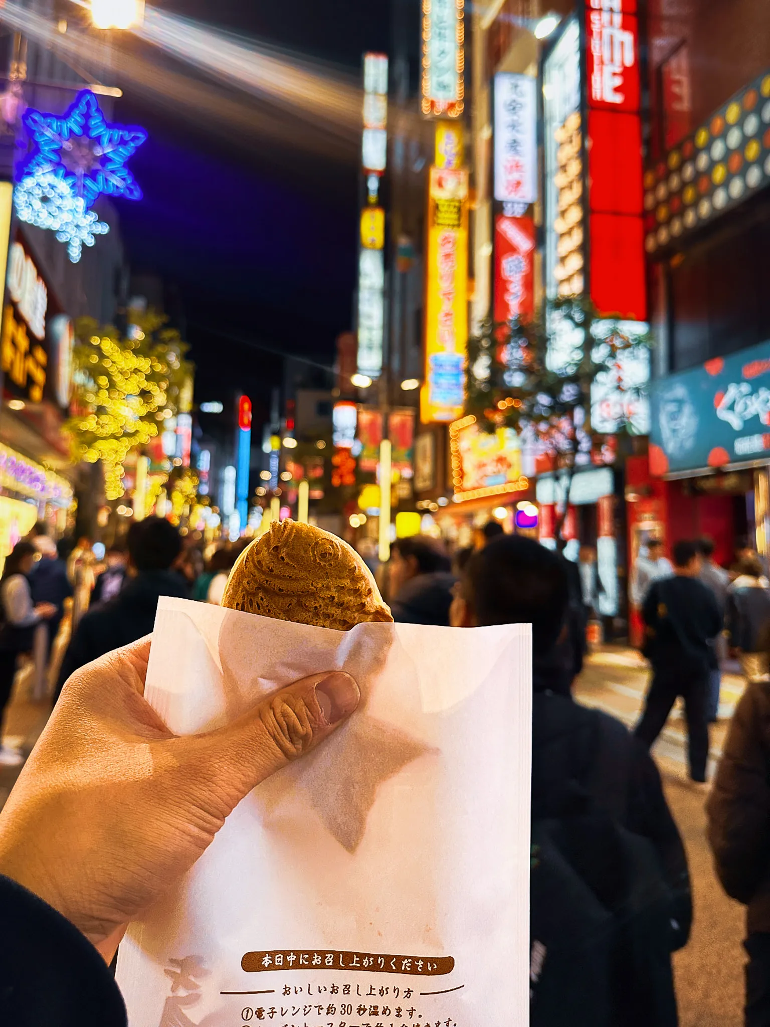A warm pastry in a paper wrapper held up against a street of neon signs and snowflake lights at night