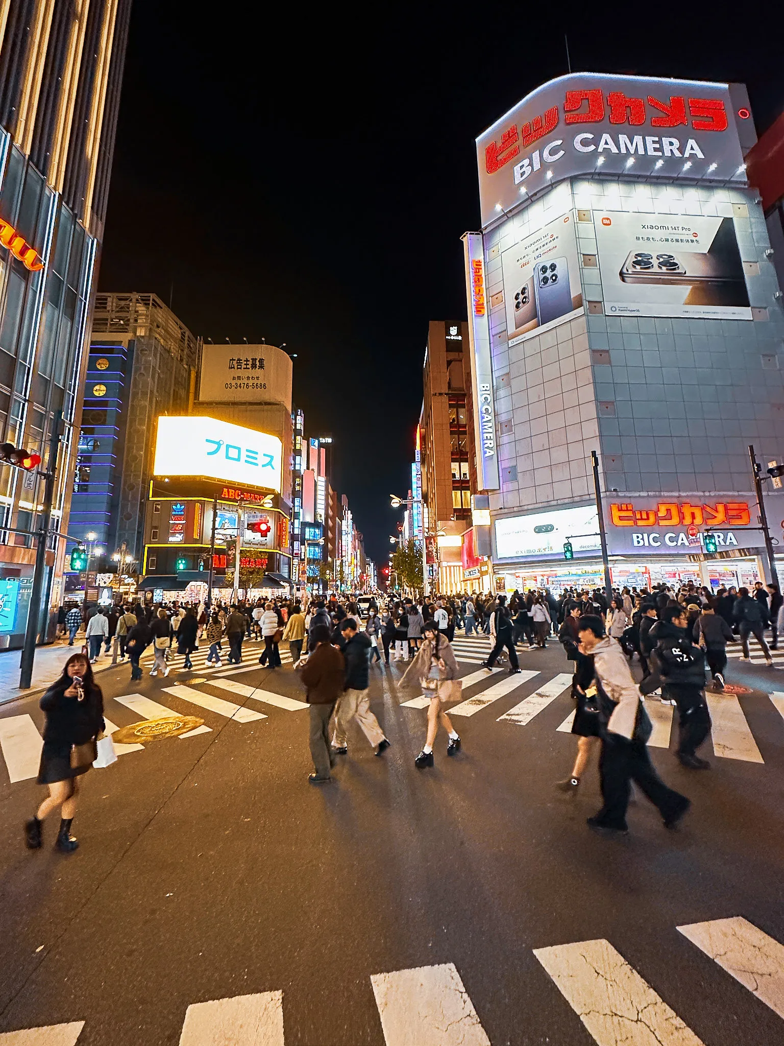 A wide crossing at night with crowds streaming across it in every direction, a camera store lit up on the right