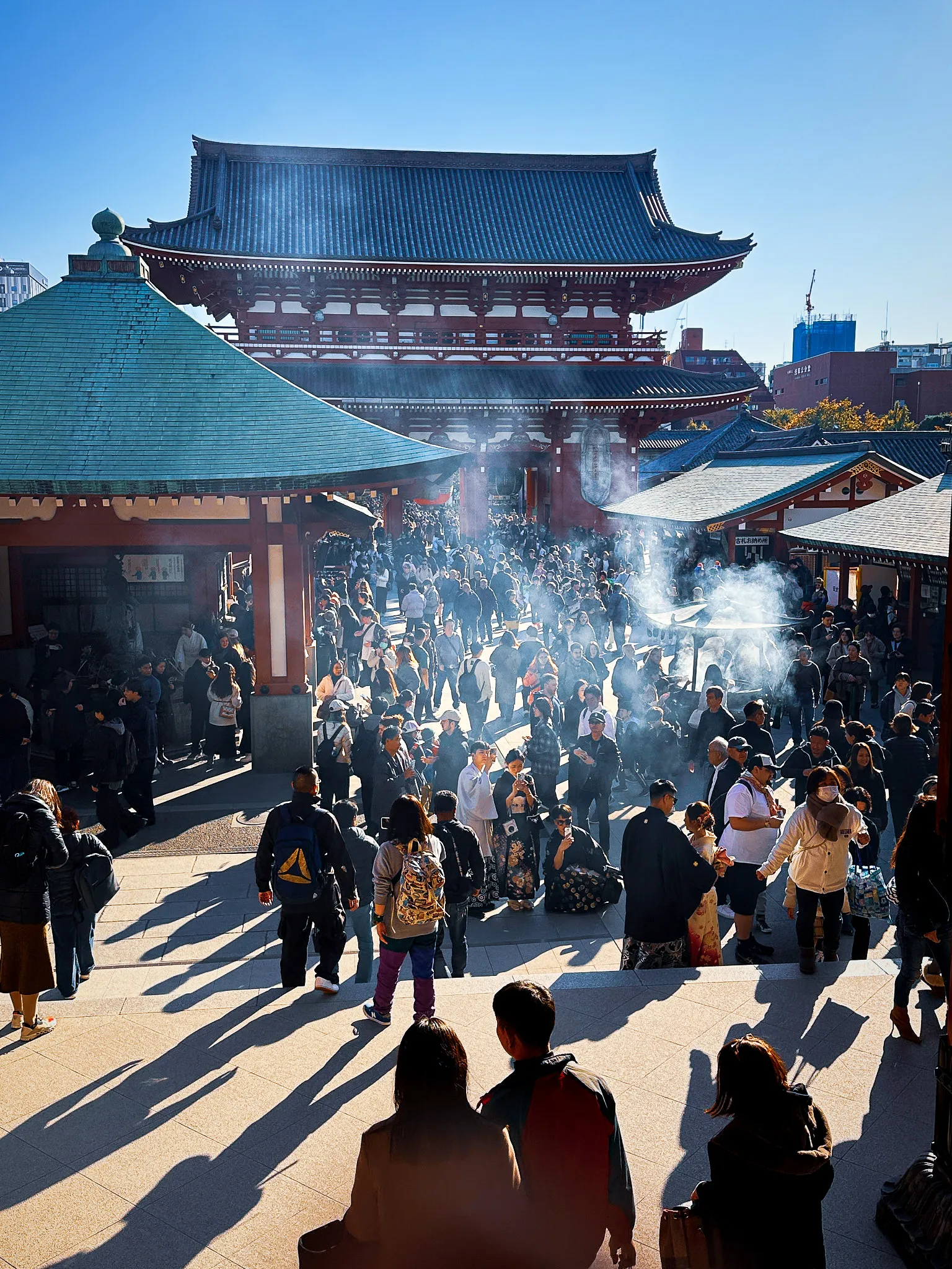 Incense smoke rising from a bronze burner in the temple courtyard, crowds gathered around it and the gate roofs behind under a blue sky