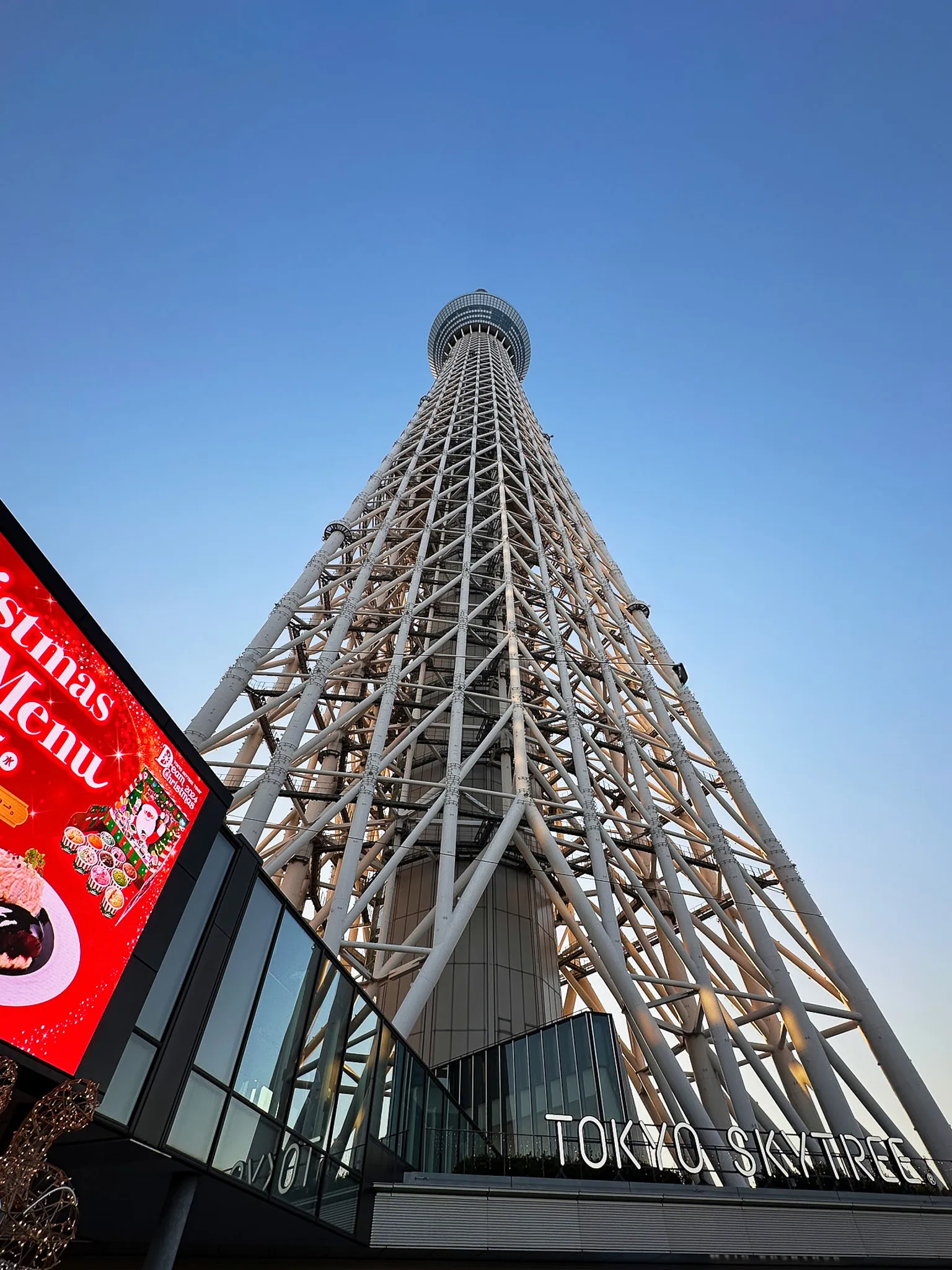 The Tokyo Skytree from its base at dusk, the white lattice tapering up into a clear blue sky, the entrance sign lit below