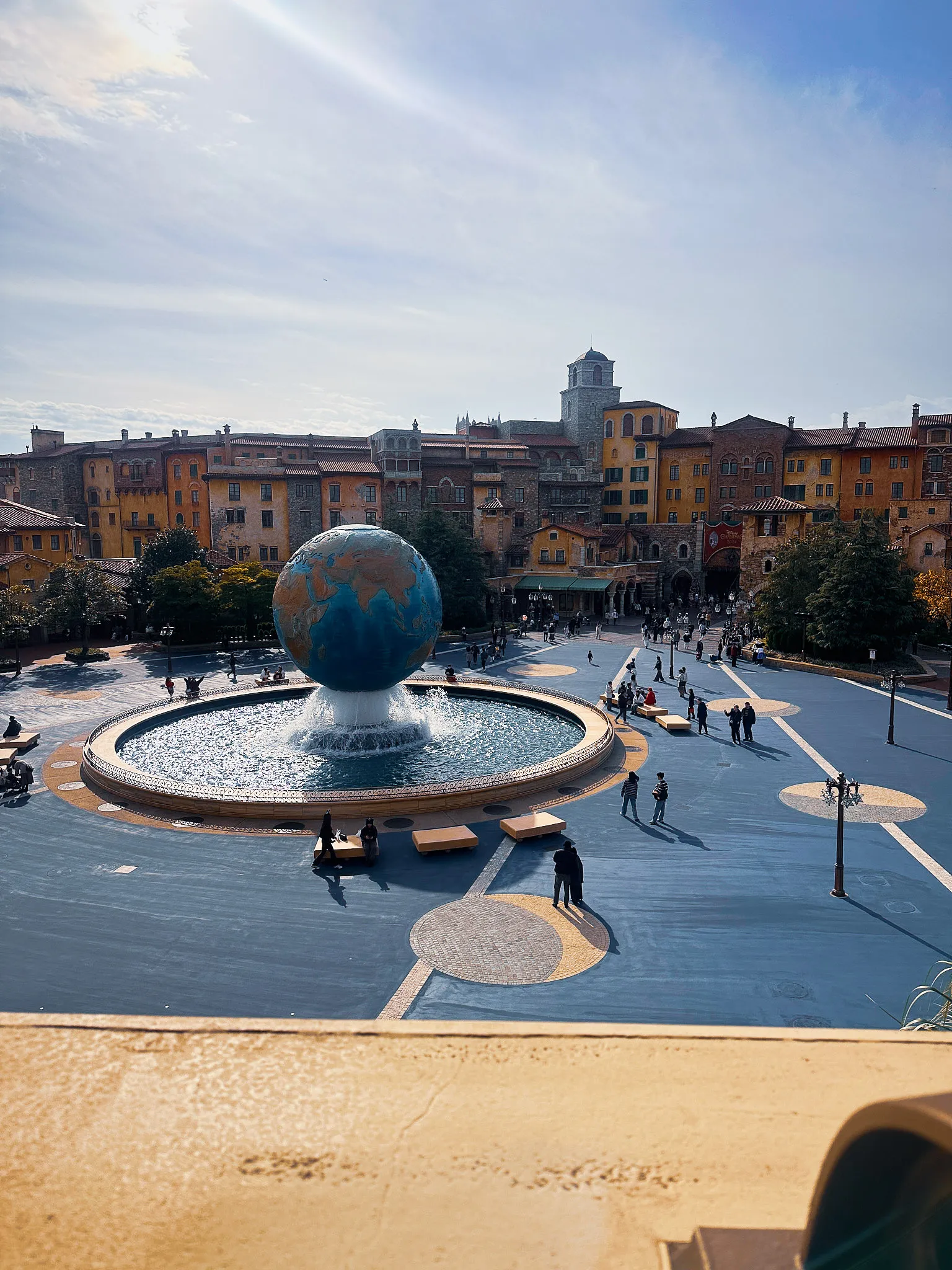 A large globe fountain in a plaza ringed by Mediterranean-style buildings, people walking the paving around it