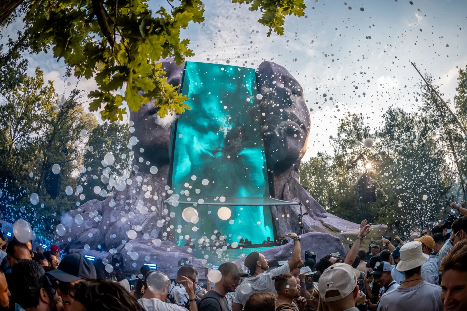 A crowd facing a turquoise-lit stage set into a sculpted rock face, confetti hanging in the trees above