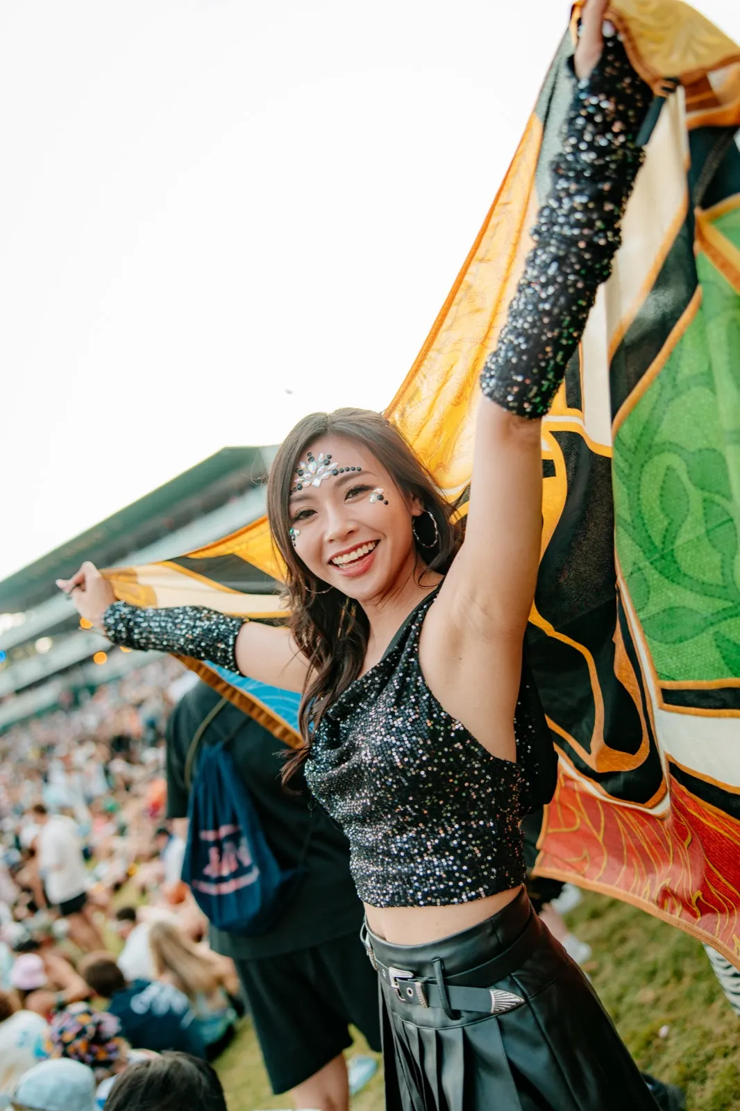 A woman lifting a flag above her head and laughing, glitter across her cheekbone, the crowd on the grass behind