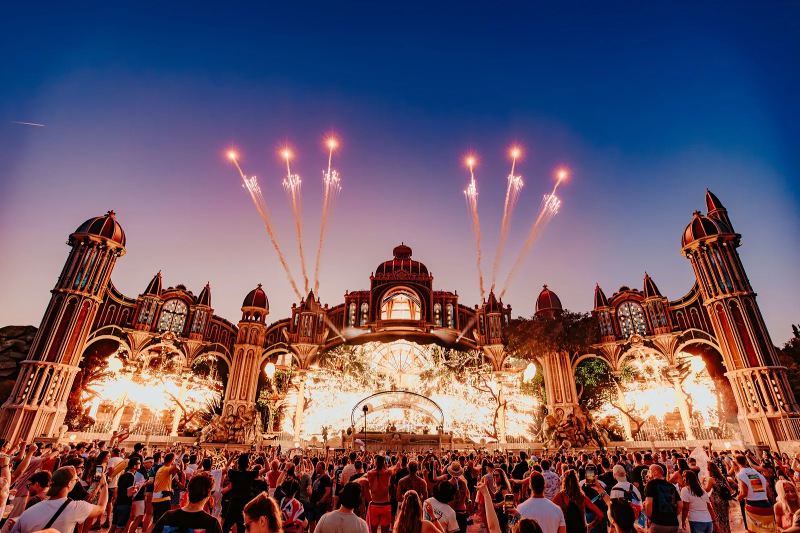 The main stage's ornate lit facade at dusk with jets of firework rising from it and a vast crowd filling the field