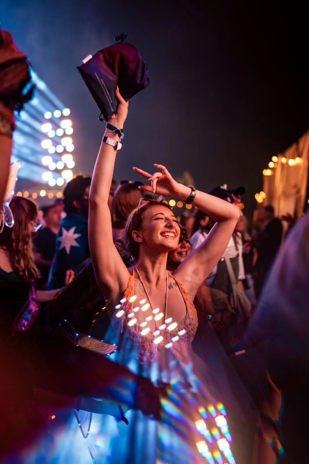 A woman in a dress covered in small lights, arms raised in the middle of the crowd under a beam of stage light
