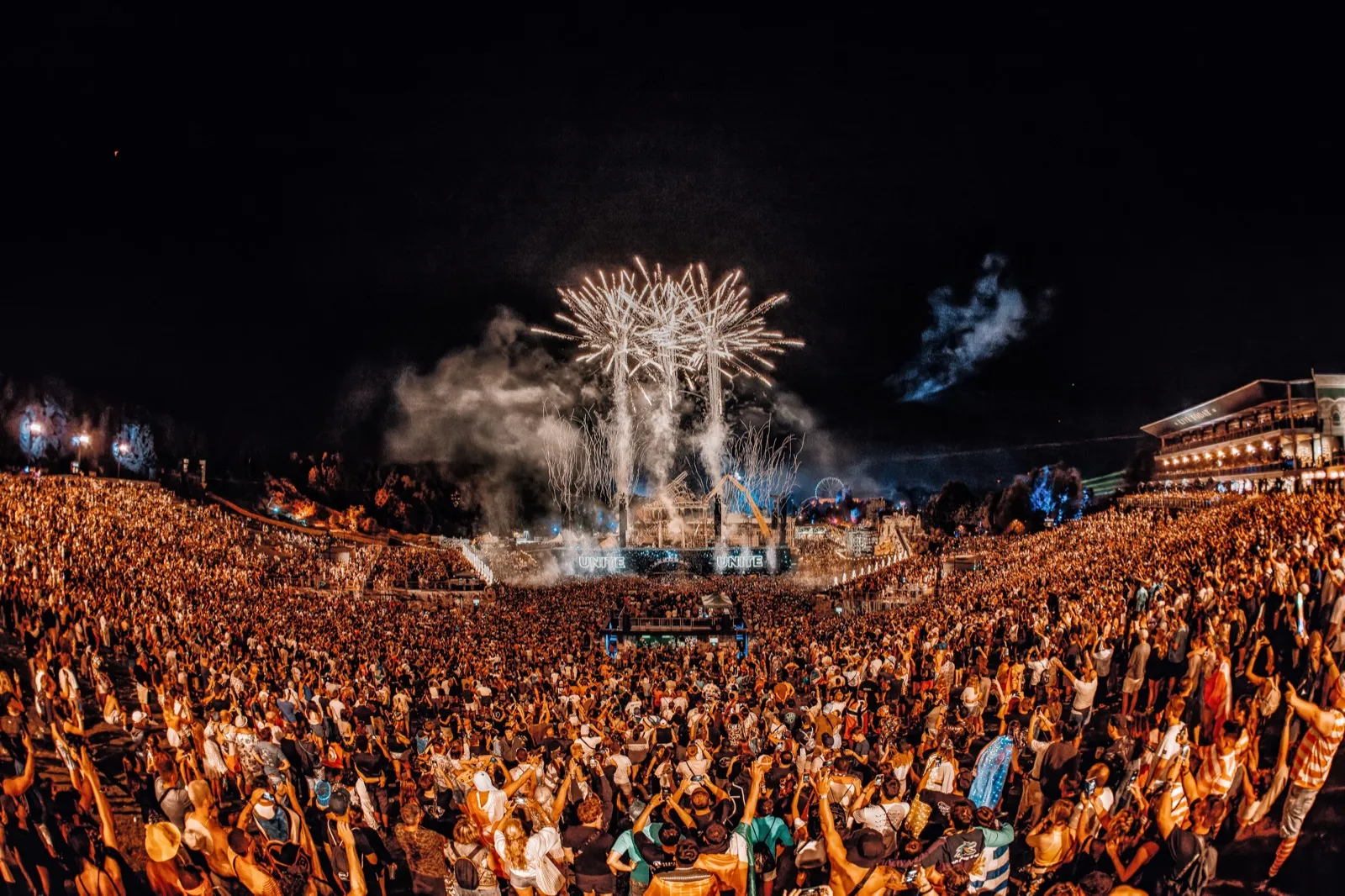 The main stage at night from far back, a burst of firework above and the crowd stretching away in every direction