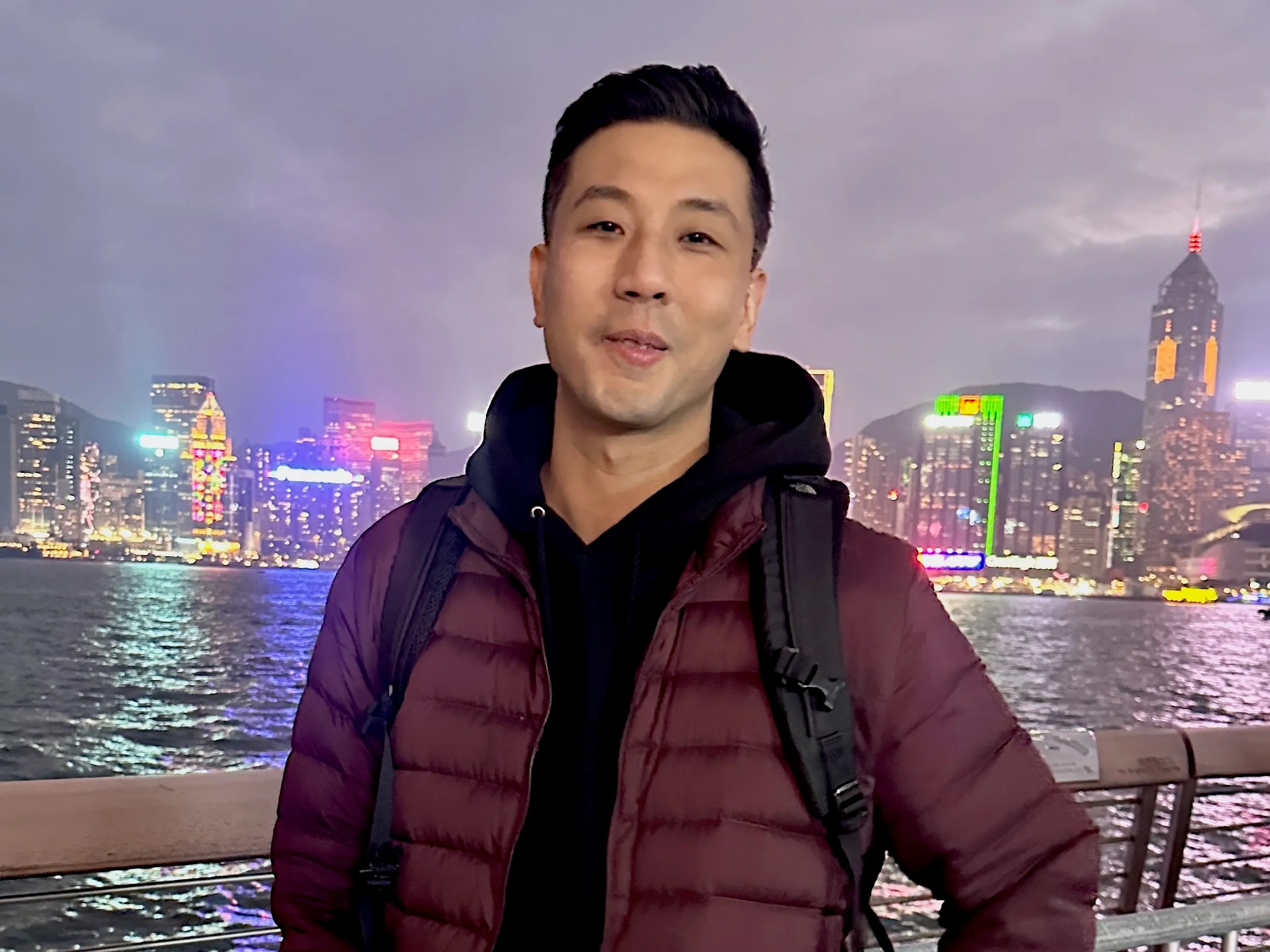 Trung at night on the waterfront, the lit skyline across the harbor behind him