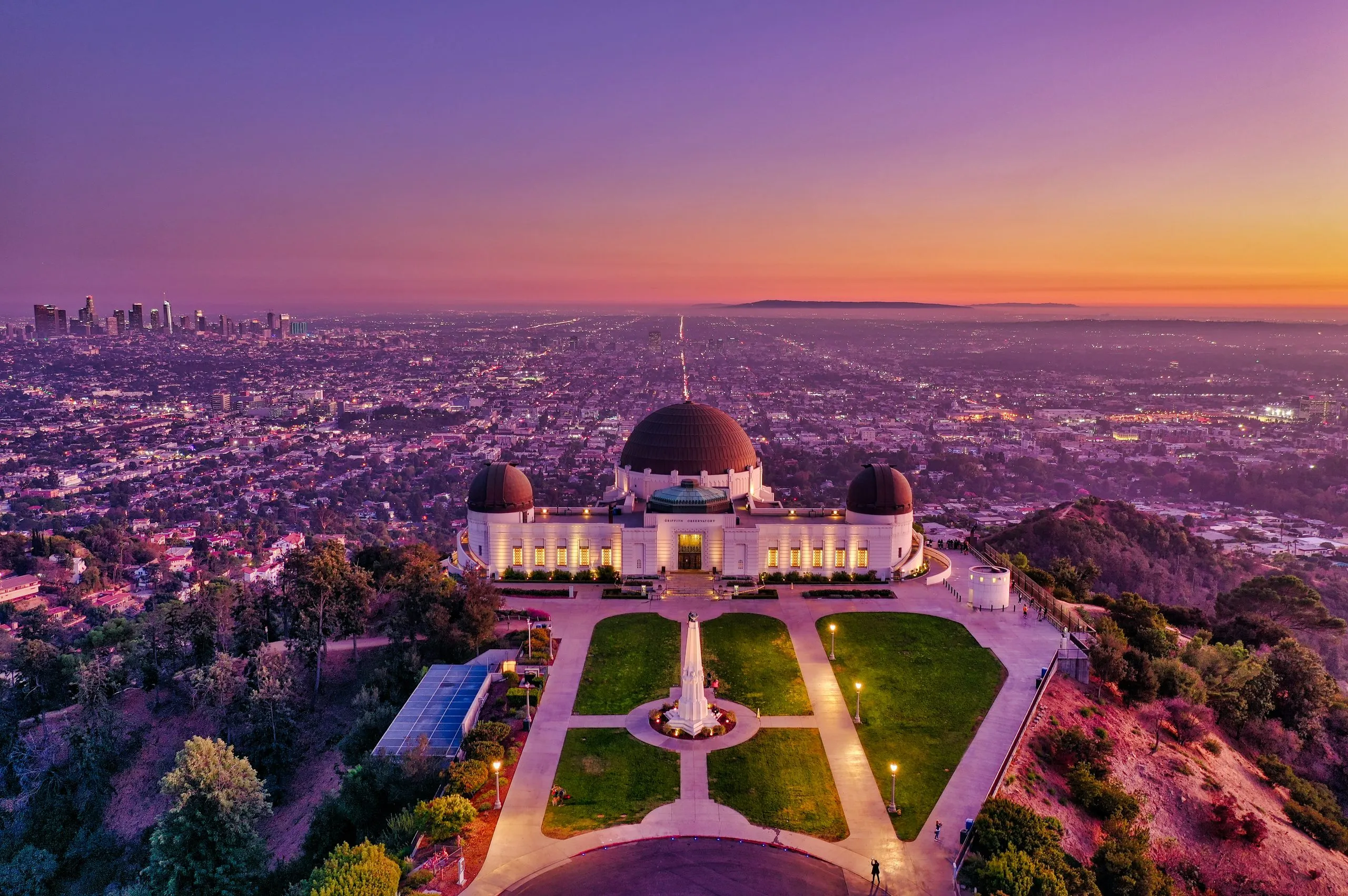 Griffith Observatory's domes from above at sunset, lawns and paths laid out in front and the city and ocean beyond in violet light